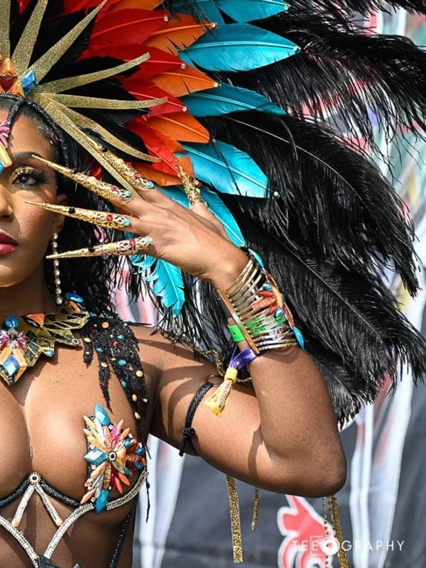 Masquerader in a blue, orange and black feathered headdress and beaded costume with long gold nails