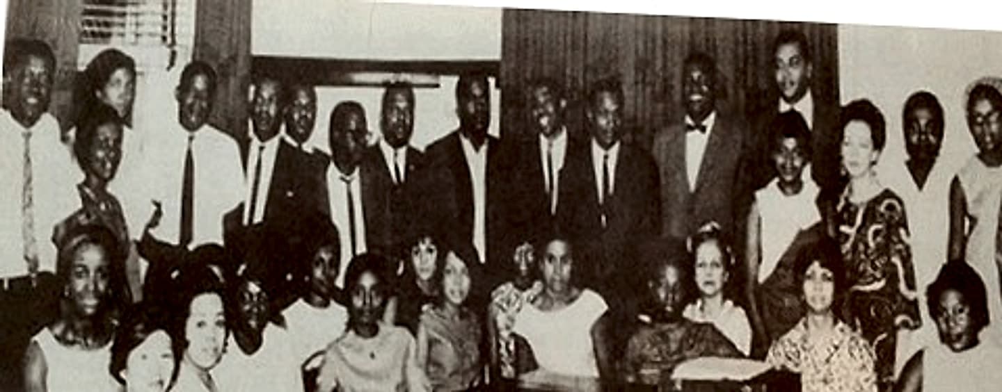 Black-and-white group photograph of about thirty men, women and young people posed in rows in a wood-panelled room