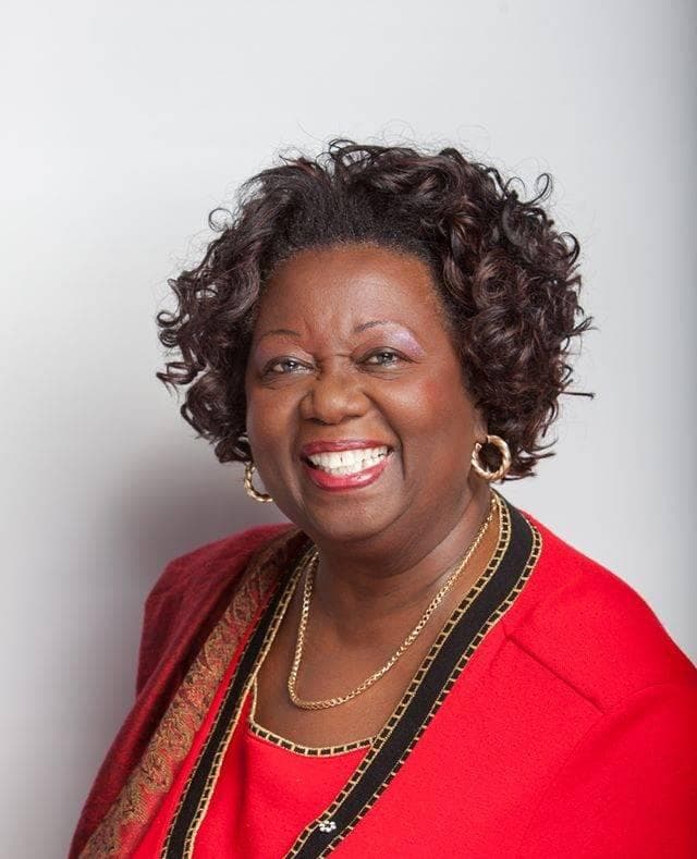 Portrait of Jean Augustine: a smiling woman with short curly hair, gold hoop earrings and a gold necklace, in a red jacket with black and gold trim, against a pale grey background