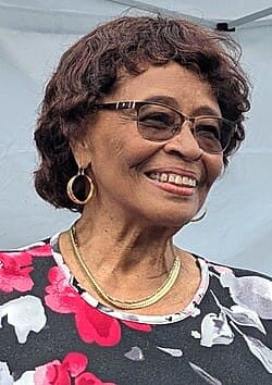 Portrait of Rita Cox: a smiling older woman with short curly hair, glasses, gold hoop earrings and a gold necklace, in a pink, black and white floral top