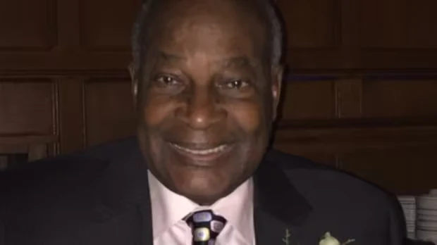 Portrait of Romain Pitt: a smiling older man in a dark suit, light shirt and patterned tie, in front of dark wood panelling