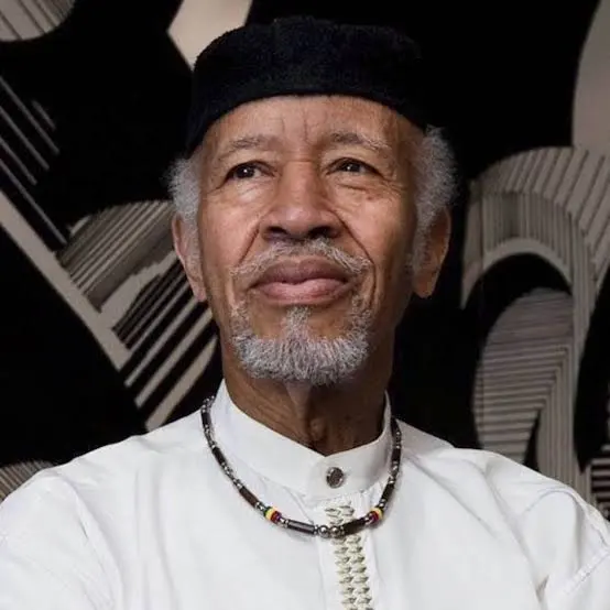 Portrait of Charles Roach: an older man with a grey beard in a black cap, white collarless shirt and a beaded necklace, in front of a black-and-white patterned wall
