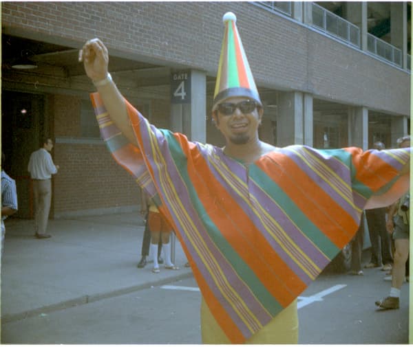 Colour photograph of Kenneth Shah: a smiling man in sunglasses and a striped cone-shaped hat, wearing a large orange, green and purple striped poncho with his arms raised, outside a brick building entrance marked Gate 4