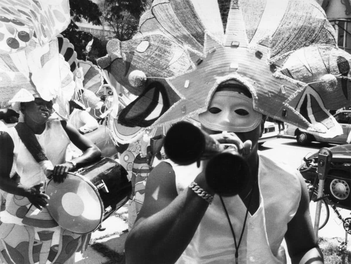 Black-and-white close-up of a masquerader in a large star-shaped headdress and white mask blowing a horn, with a drummer behind