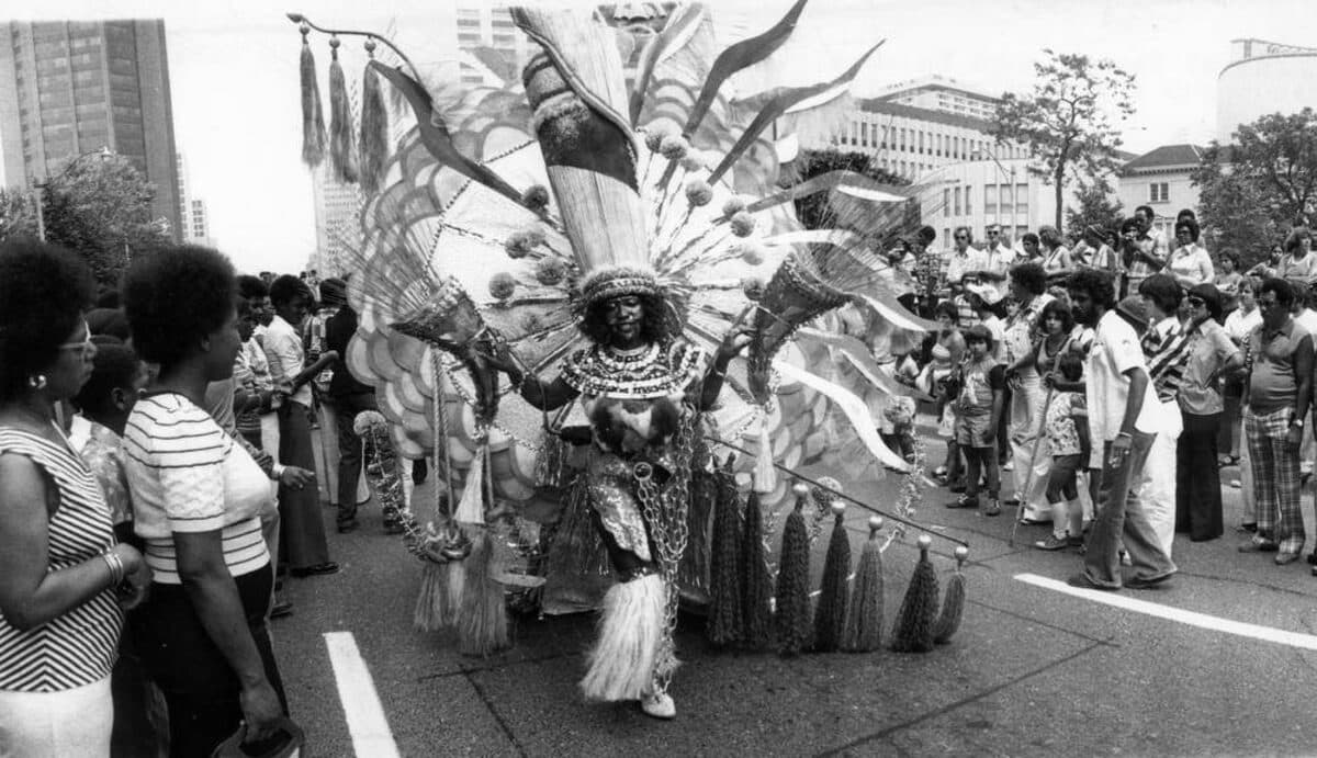 Black-and-white photograph of a masquerader in a towering feathered costume with crowds lining a street between tall buildings
