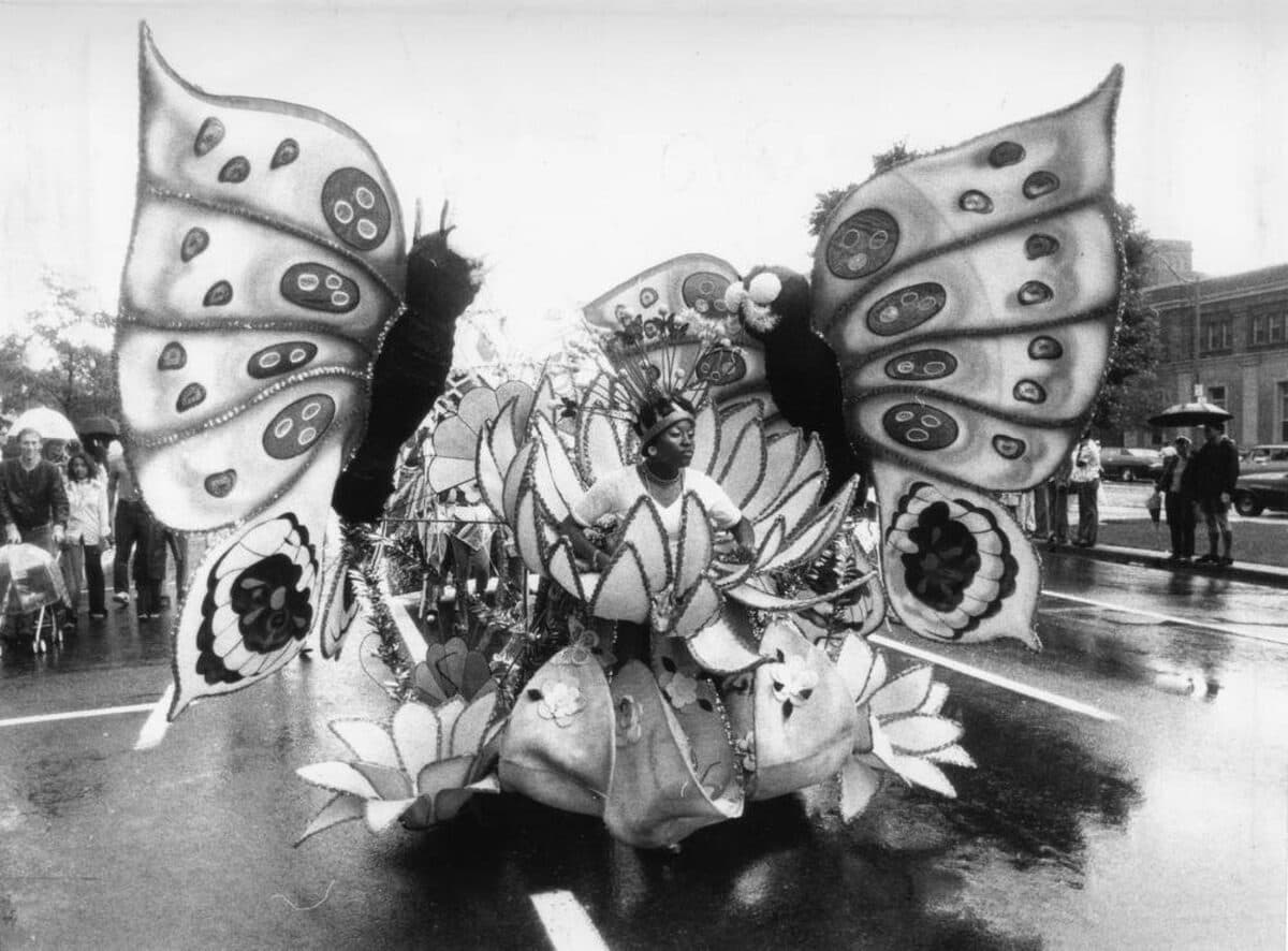 Black-and-white photograph of a masquerader in a large butterfly costume with spotted wings riding a float along a wet street in the rain