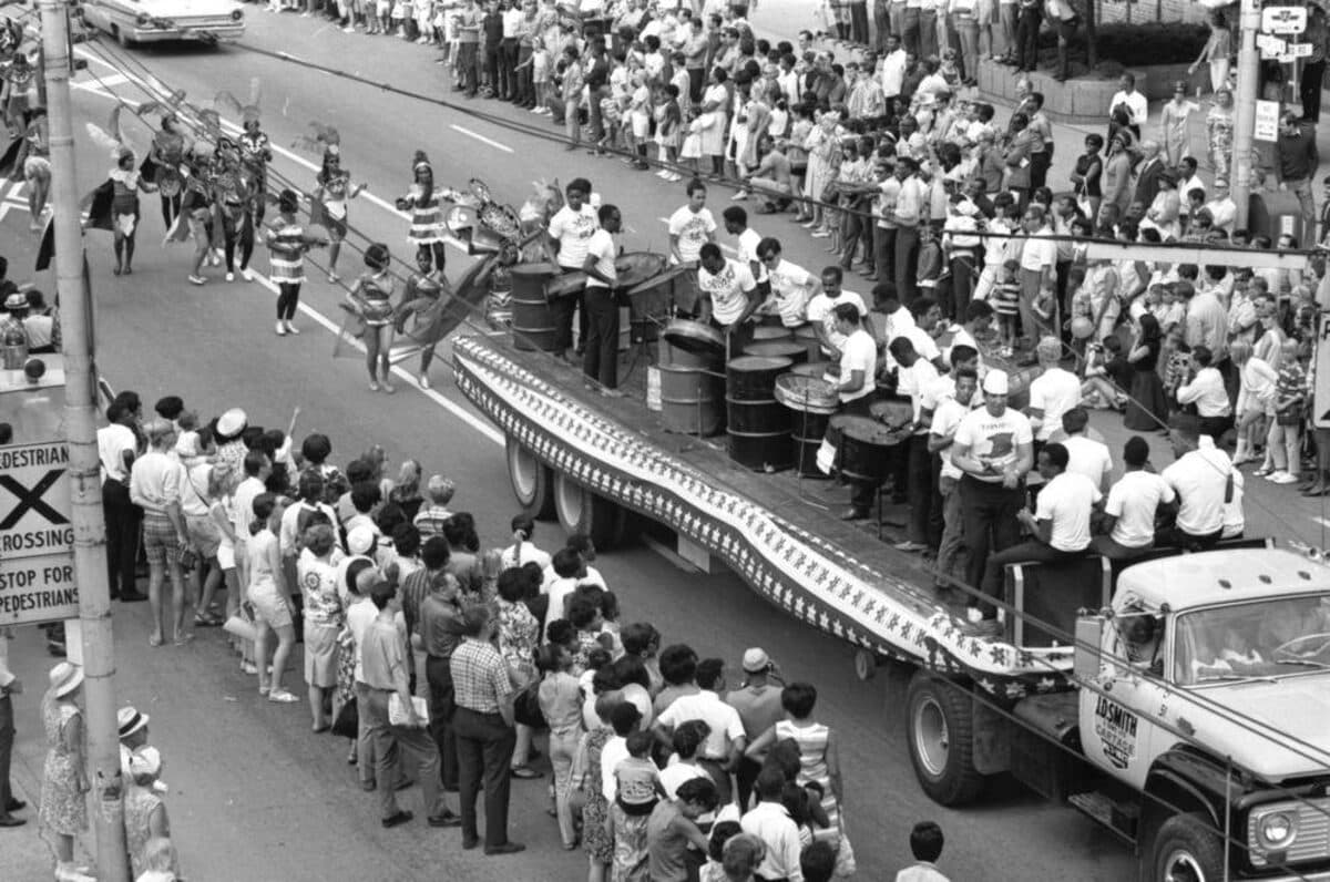 Black-and-white photograph taken from above of musicians playing drums on a flatbed truck in a street parade, with crowds along the road