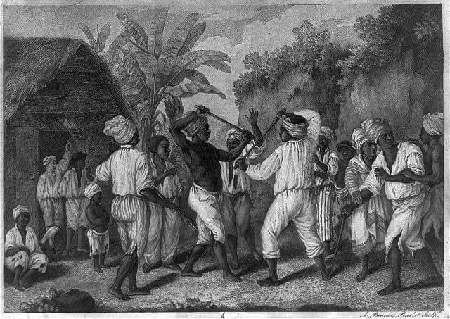 Black-and-white engraving of men in turbans and loose white clothes fighting with sticks in front of a thatched hut, watched by onlookers under banana trees