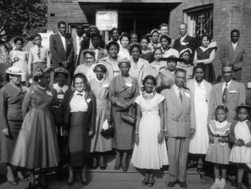 Black-and-white group photograph of about thirty women, men and children in smart clothes standing in rows on the steps of a brick building