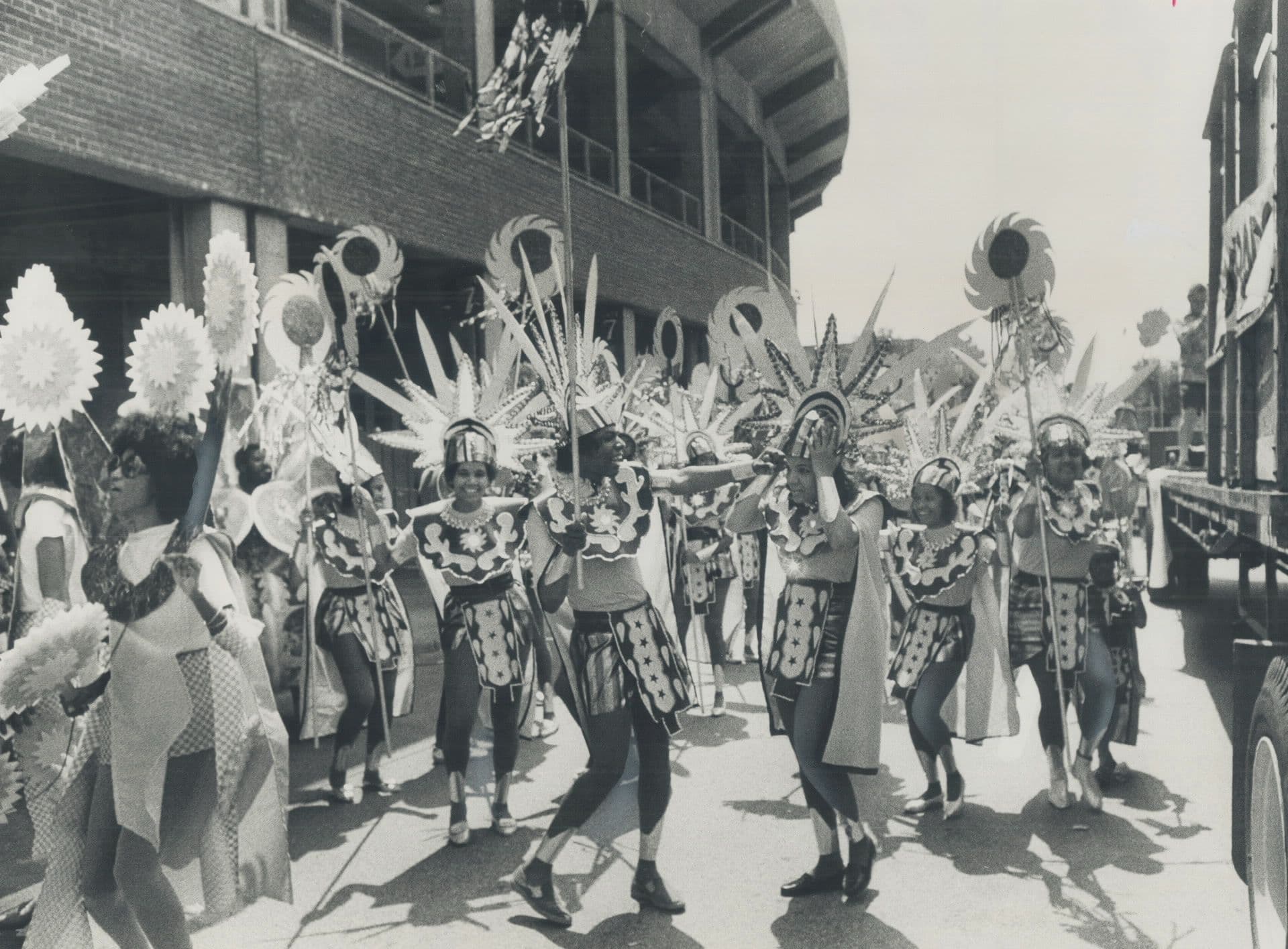 Black-and-white photograph of masqueraders in sun-shaped headdresses and patterned costumes dancing along a street beside a stadium